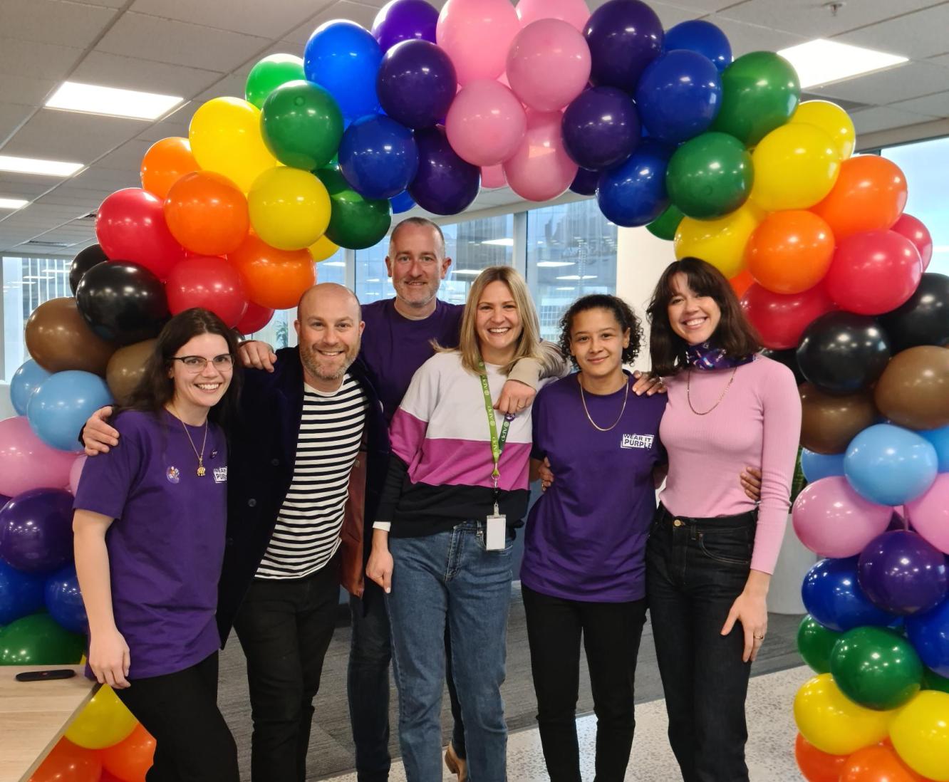 Six colleagues standard under a rainbow coloured balloon arch